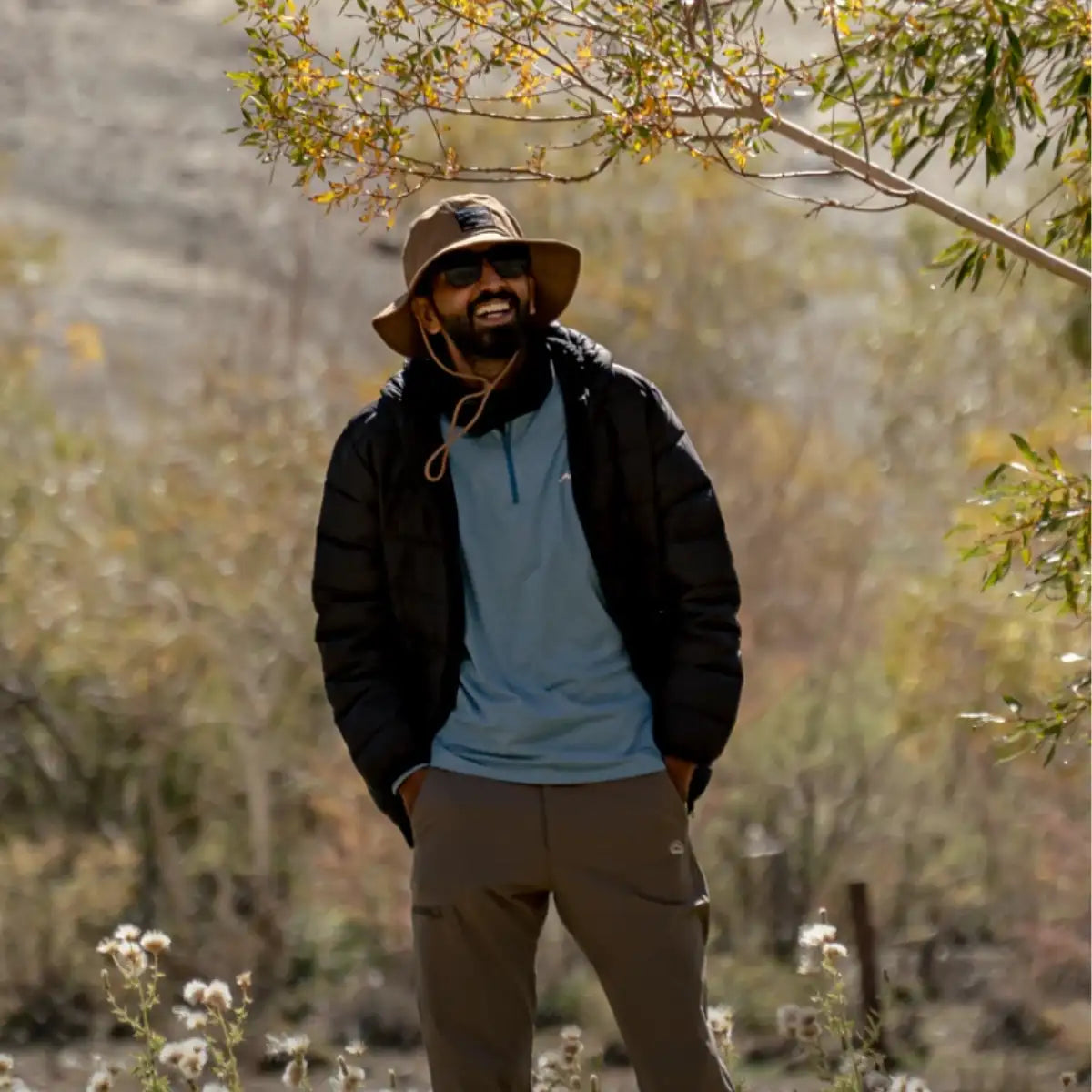 A man wearing a brown wide-brimmed hat, black puffer jacket over a light blue shirt, and khaki pants stands outdoors under tree branches.