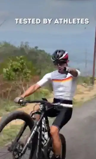 A cyclist wearing a white jersey with orange and black accents, black shorts, and a helmet is seated on a mountain bike.