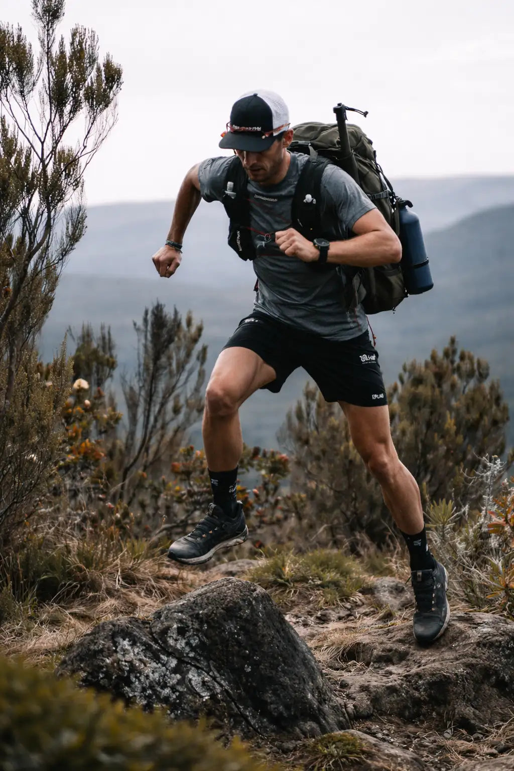 A man wearing a gray and black athletic outfit with a white-and-black cap, carrying a large olive green backpack equipped with gear.