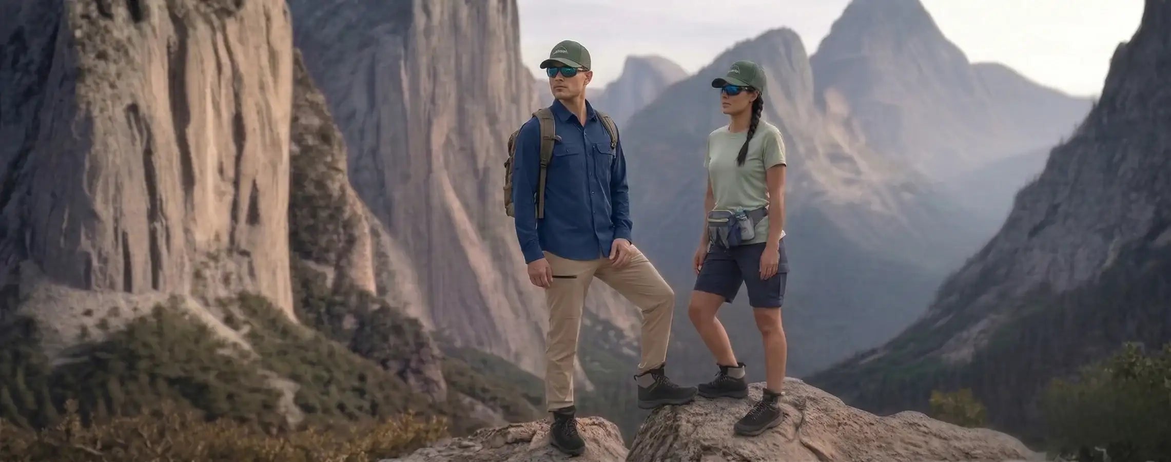 A pair of hikers stand on a rocky outcrop, dressed in matching green caps and outdoor gear against the backdrop of towering granite cliffs.