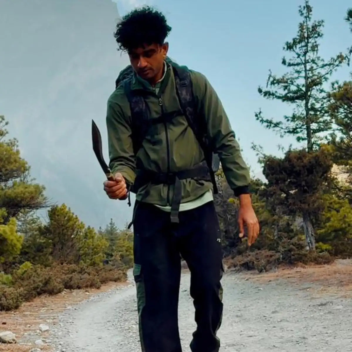 A person wearing a green jacket and black pants, carrying a backpack while holding a curved-bladed knife.