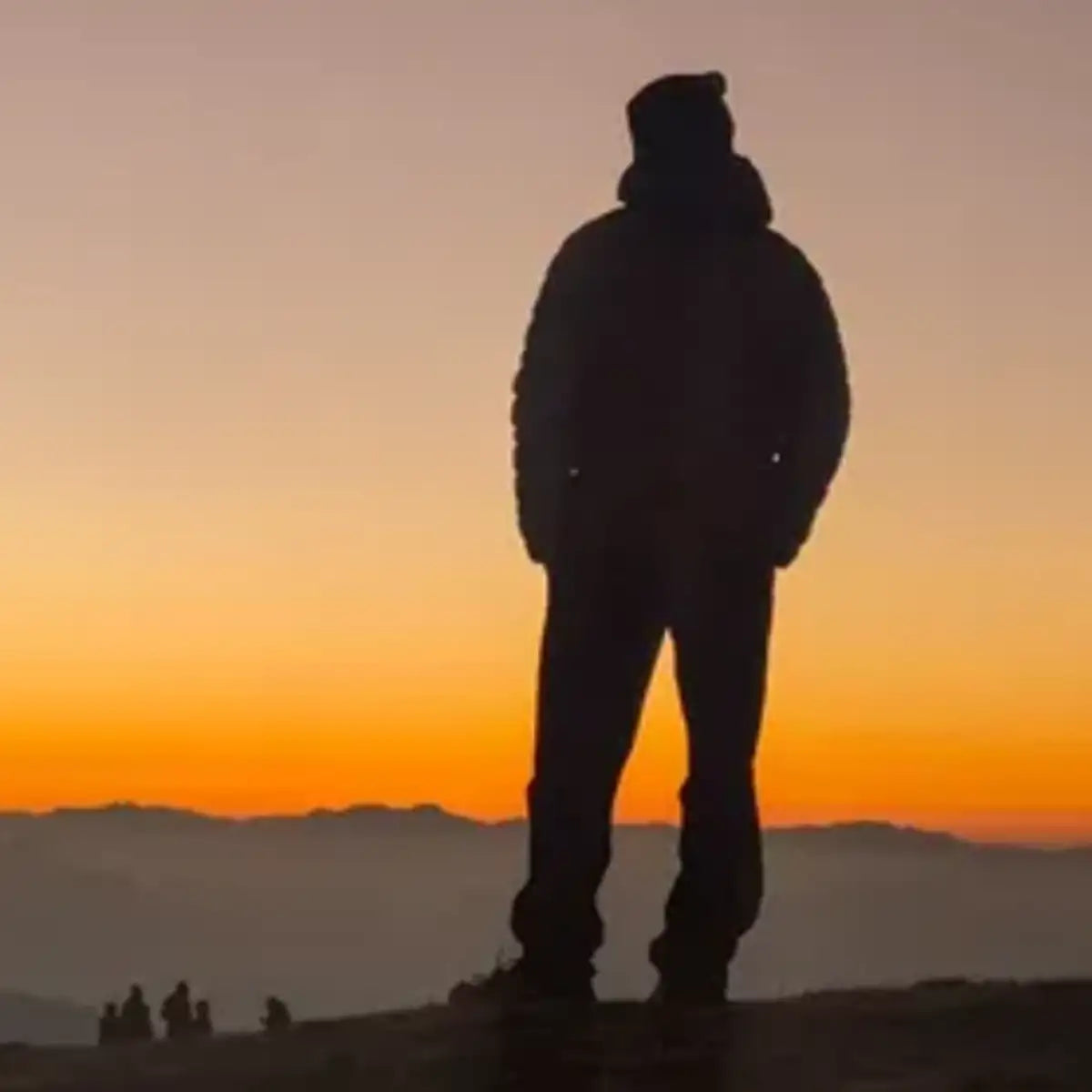Silhouetted figure standing atop a mountain at sunset, wearing a hooded jacket and beanie.