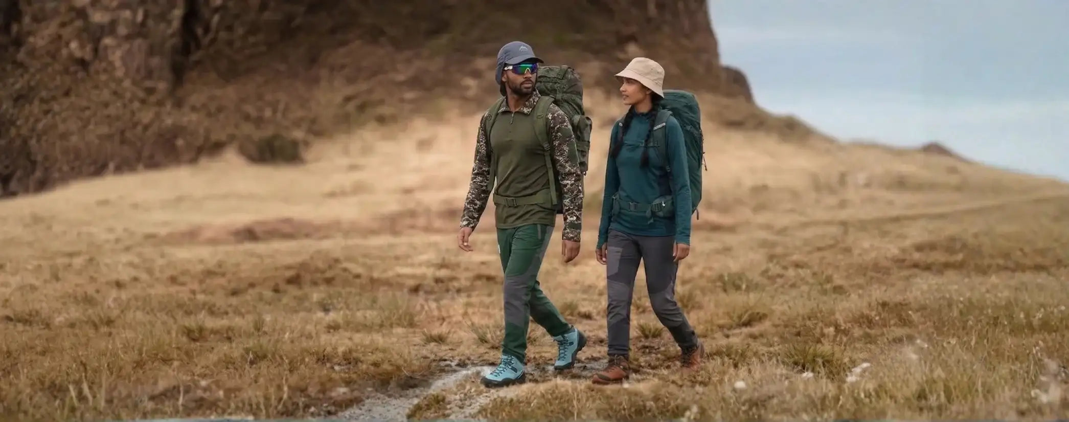 Two hikers, one in a camouflage-patterned long-sleeve shirt and green pants with blue hiking boots, the other in a teal jacket and dark gray trousers with brown hiking shoes.